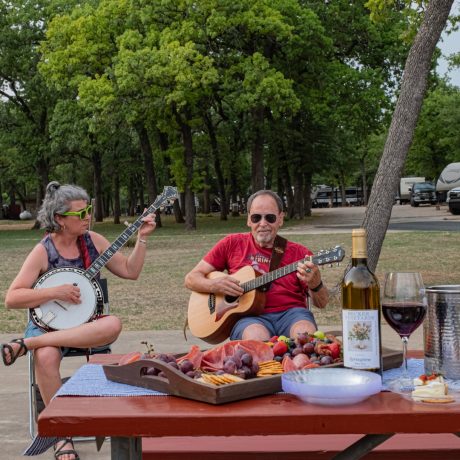 A happy friends singing and playing instruments at Oakwood RV Resort in Fredericksburg, TX, surrounded by beautiful shady views and rustic charm.